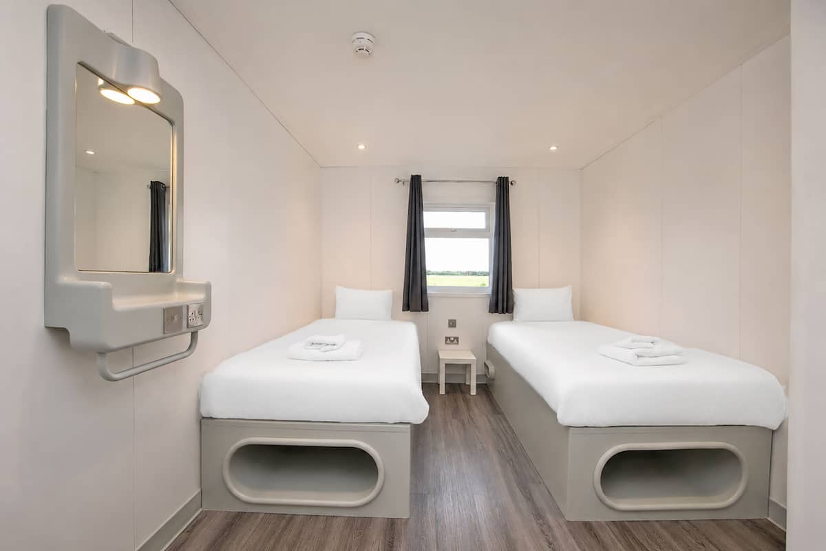 Quarto de hotel com duas camas, uma mesinha, um espelho e uma janela com cortinas.