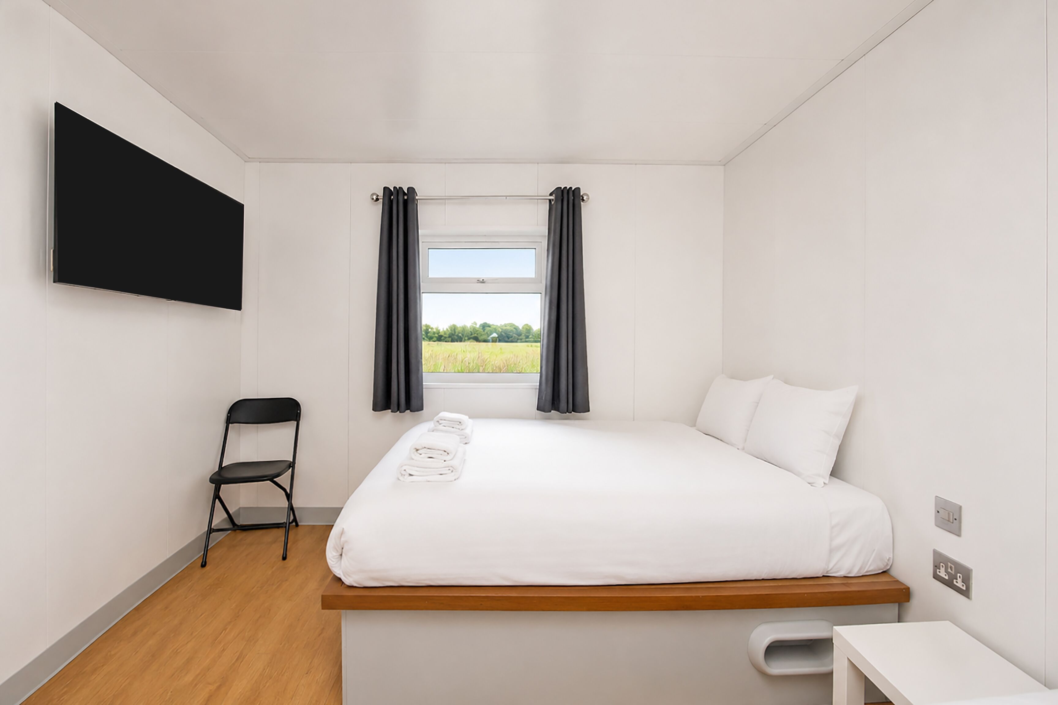 Un dormitorio con cama, una silla, una mesita, un televisor y una ventana con cortinas.