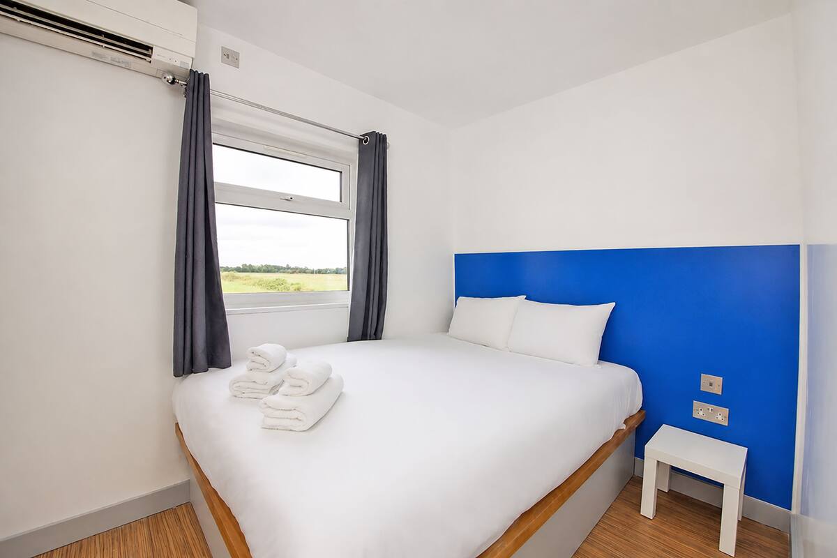 Quarto de hotel com uma cama, uma mesinha de cabeceira, uma janela com cortinas e vista para o exterior.
