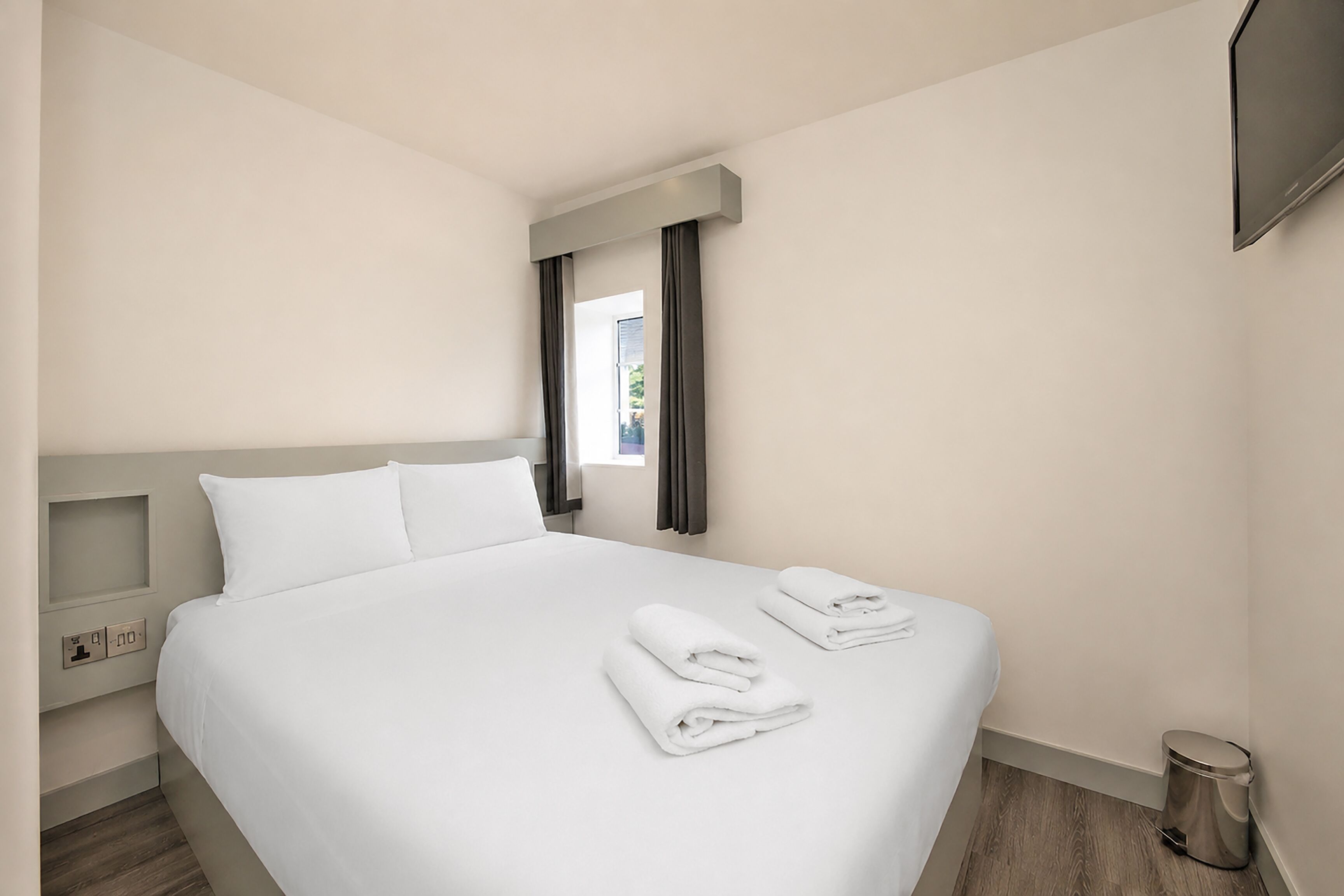 A neatly made bed with white linens, a small bedside table, a wall-mounted TV, and a window with curtains.