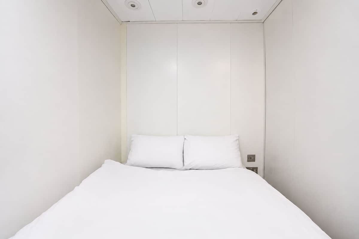 Quarto branco com uma cama de casal, parede e teto brancos, uma mesinha de cabeceira e uma tomada.