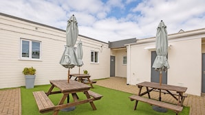 Eine Terrasse mit Picknicktischen und Sonnenschirmen, ein Pflasterweg und ein Gebäude mit Flachdach.