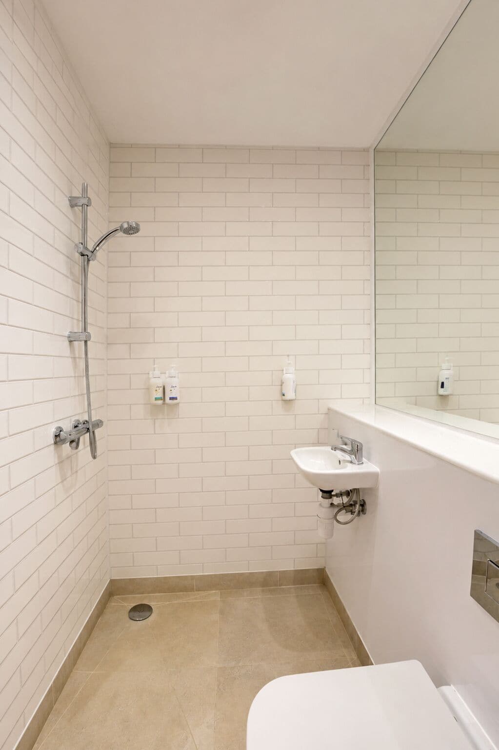 Un baño pequeño con azulejos blancos, que incluye una ducha, un lavamanos y un inodoro.