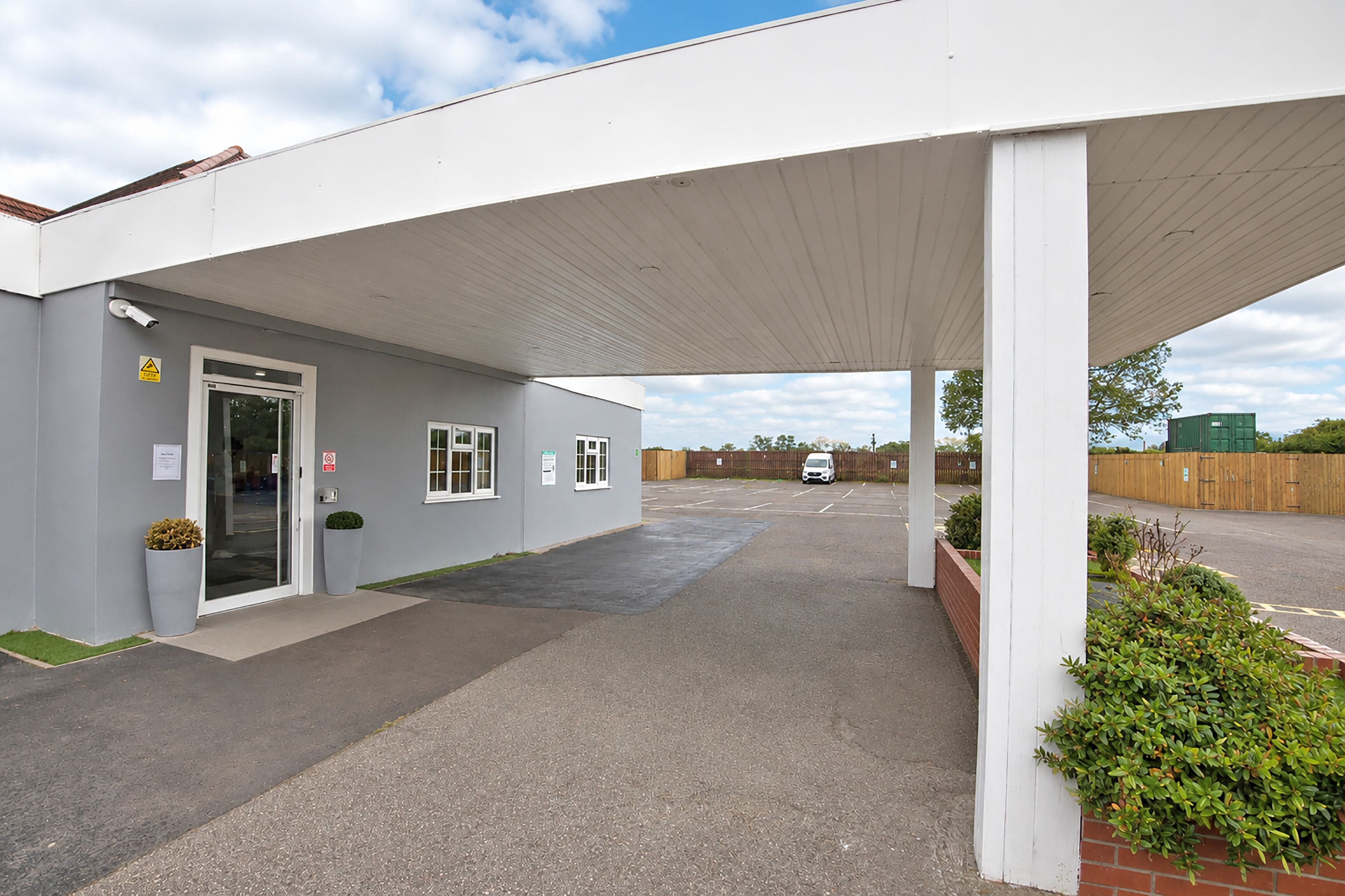 A building with a covered entrance, a parking area, and a fenced perimeter.