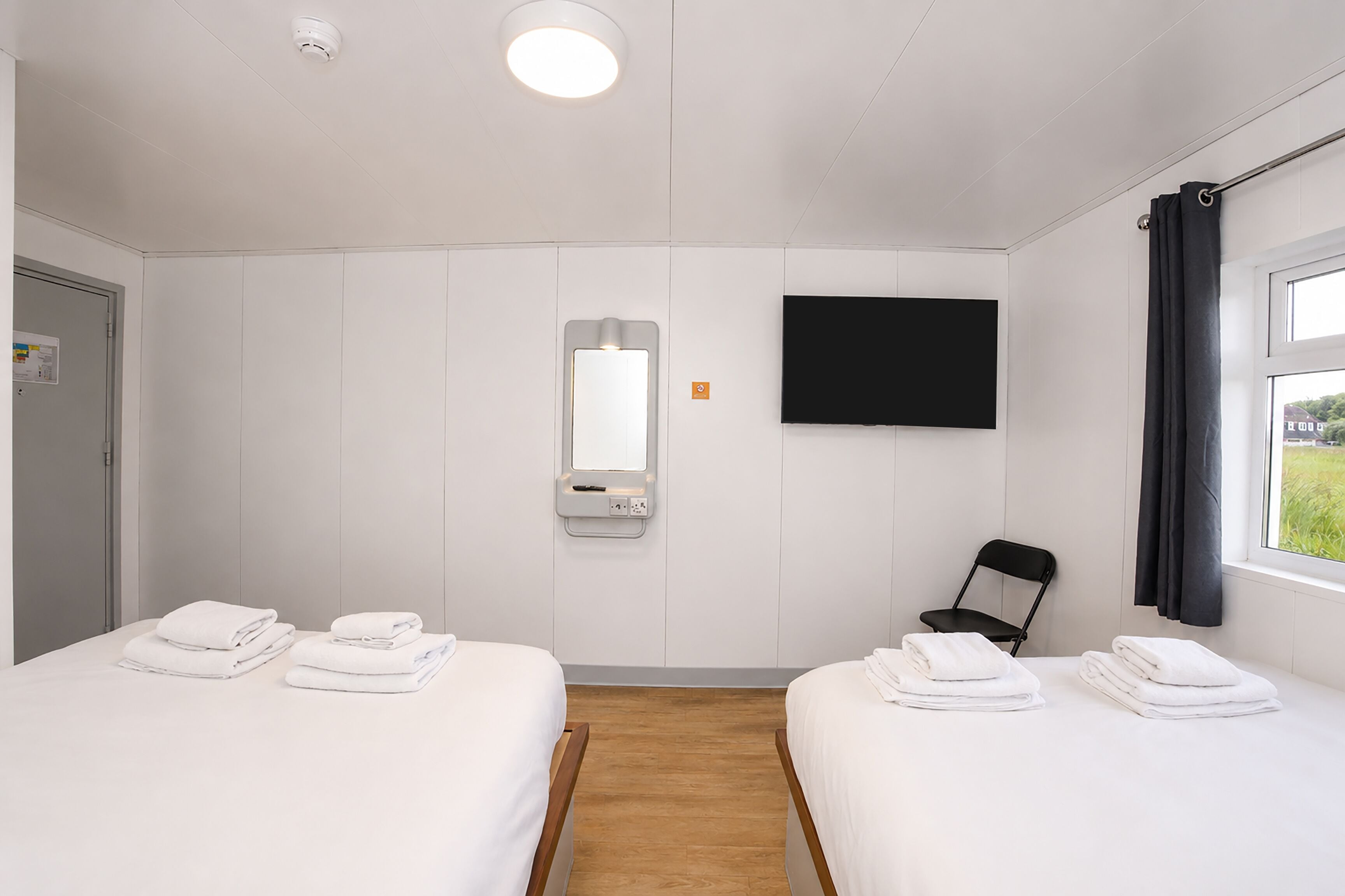 A hotel room with two beds, a TV, a mirror, and a window with curtains.