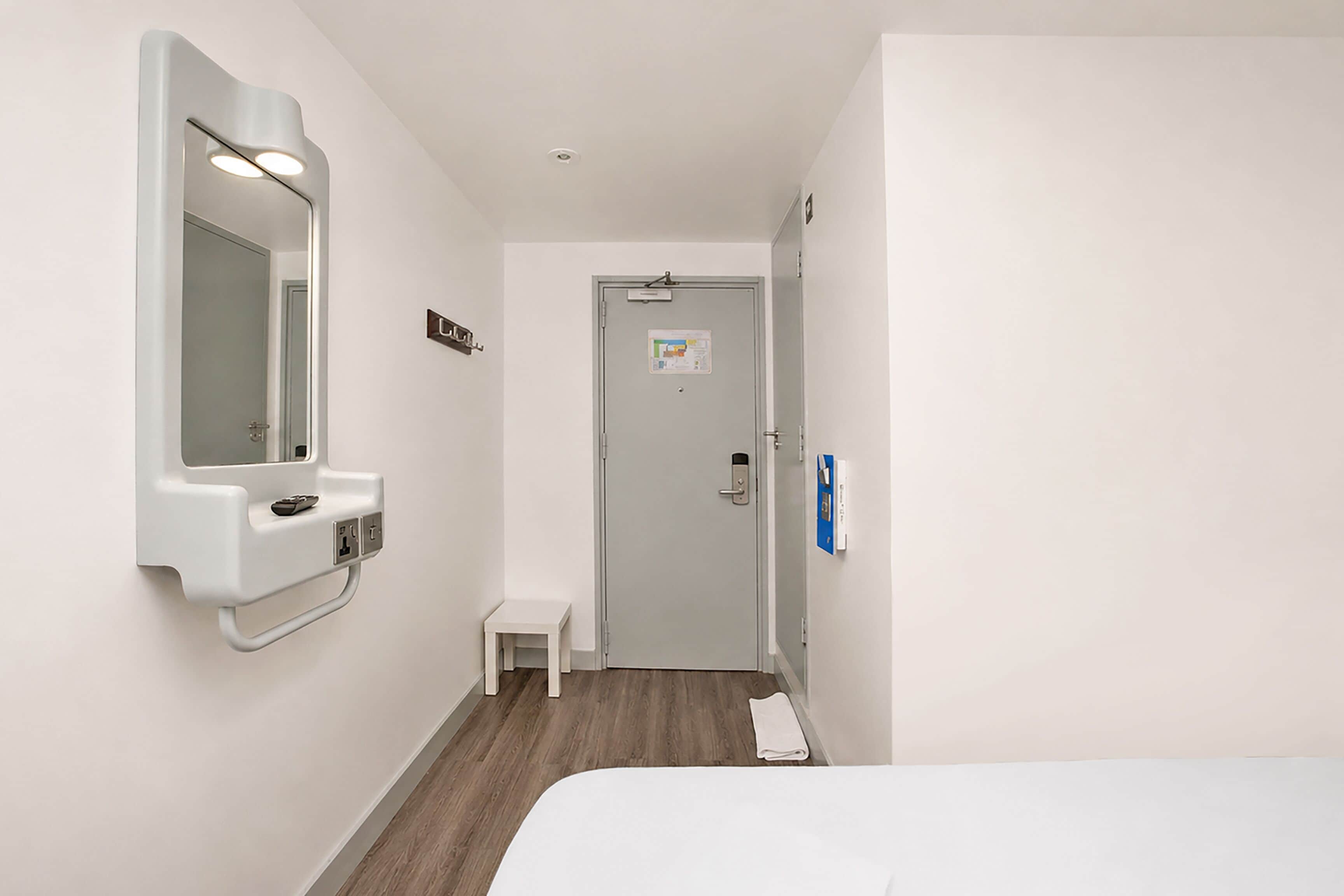 A hotel room with a bed, a small bedside table, a mirror, a door with a lock, and a wall-mounted light fixture.