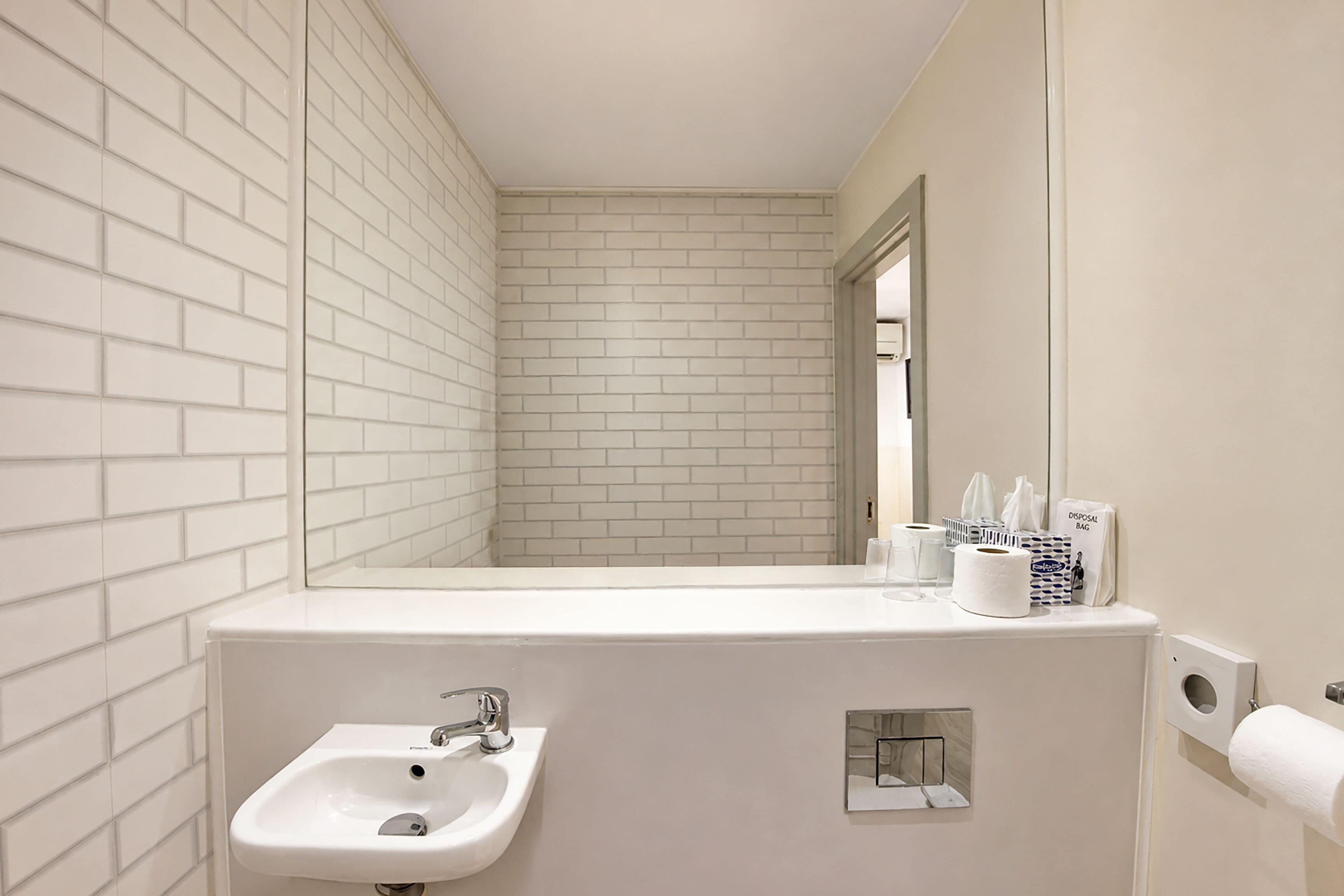 A bathroom with white tiled walls, a sink, a mirror, and a toilet paper holder.