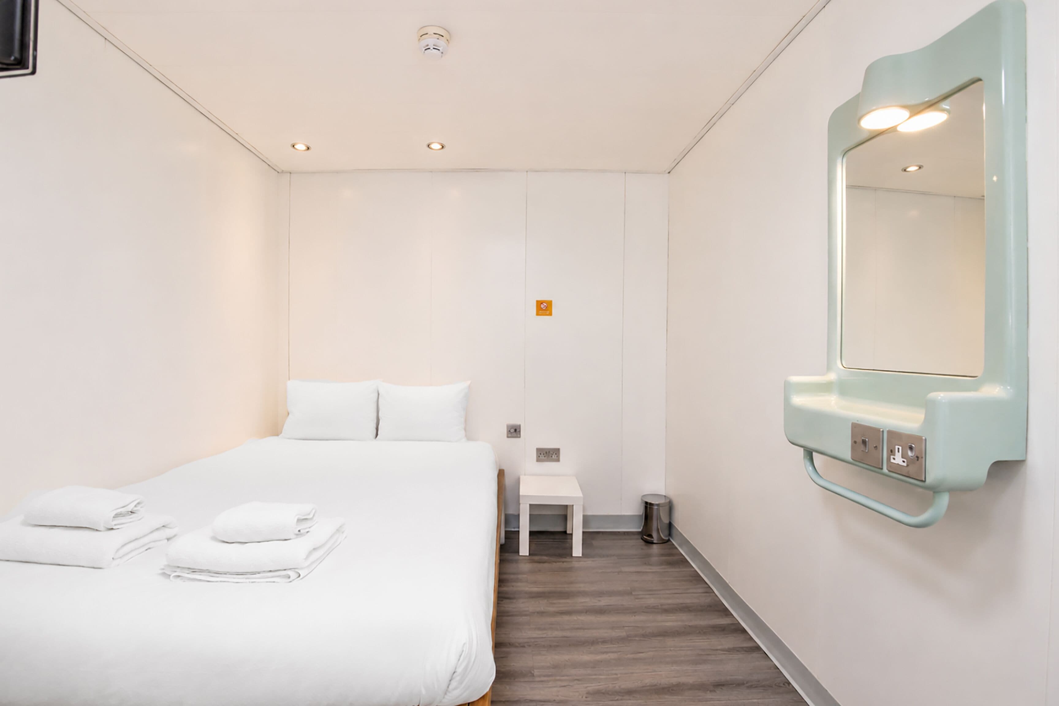Une chambre d’hôtel avec un lit, une petite table de chevet, un miroir et une poubelle.