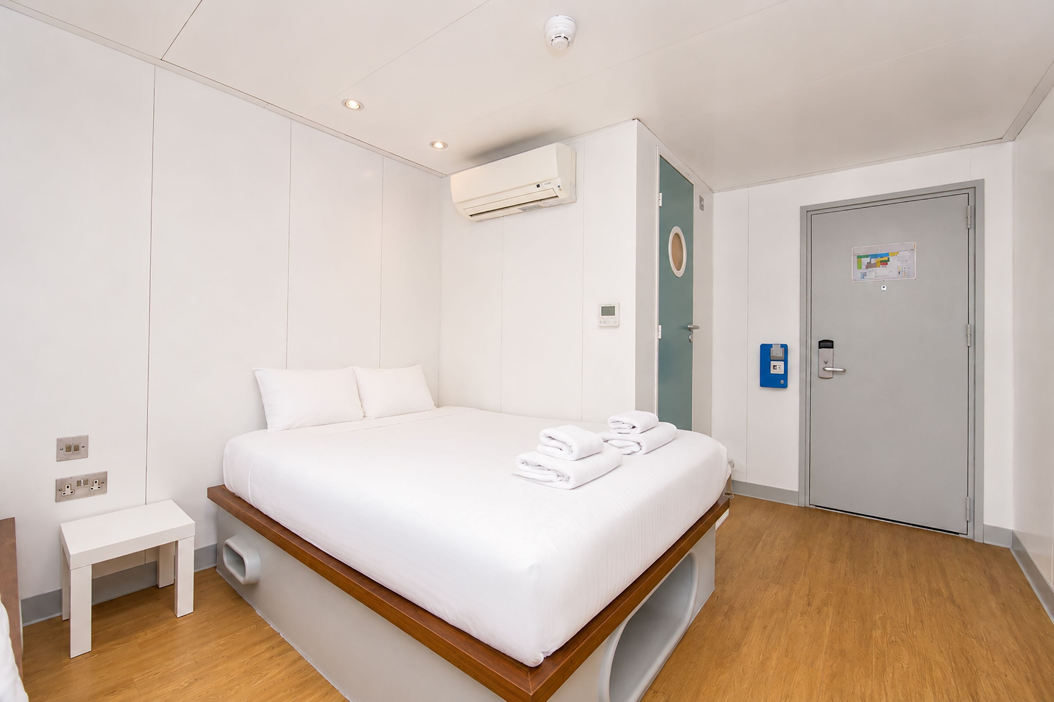 A hotel room with a bed, bedside table, air conditioning unit, door, and a small wall-mounted control panel.