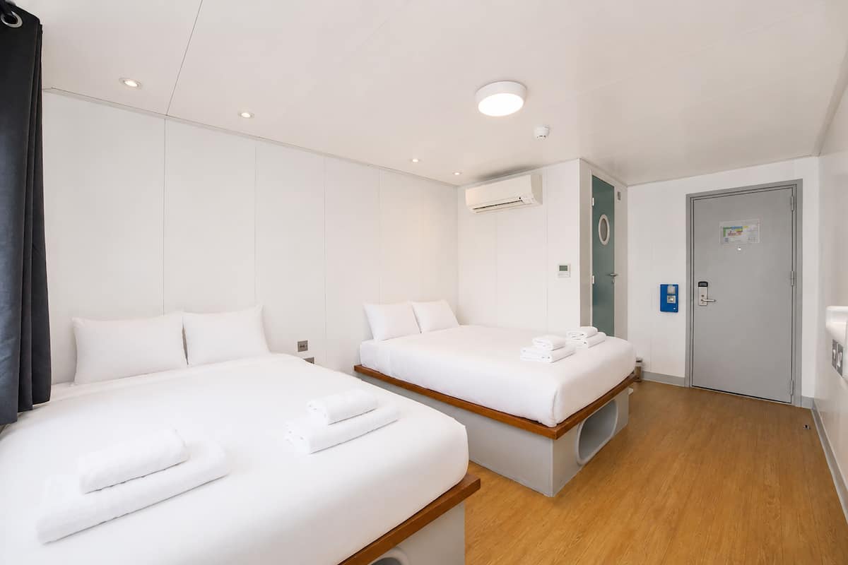 Quarto de hotel com duas camas, piso de madeira, unidade de ar condicionado, porta com fechadura e uma pequena lixeira.