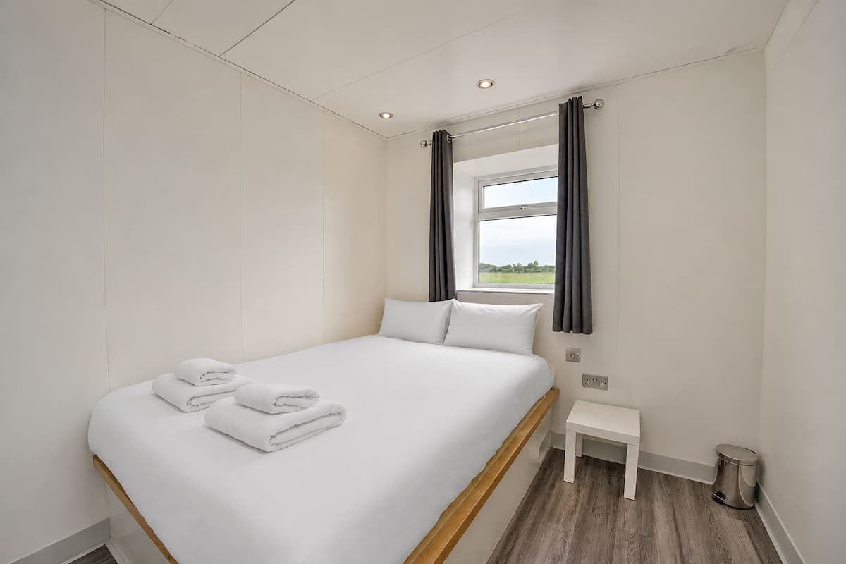 Cama bem arrumada com lençóis e toalhas brancas, uma pequena mesa de cabeceira, uma janela com cortinas e vista para o exterior.
