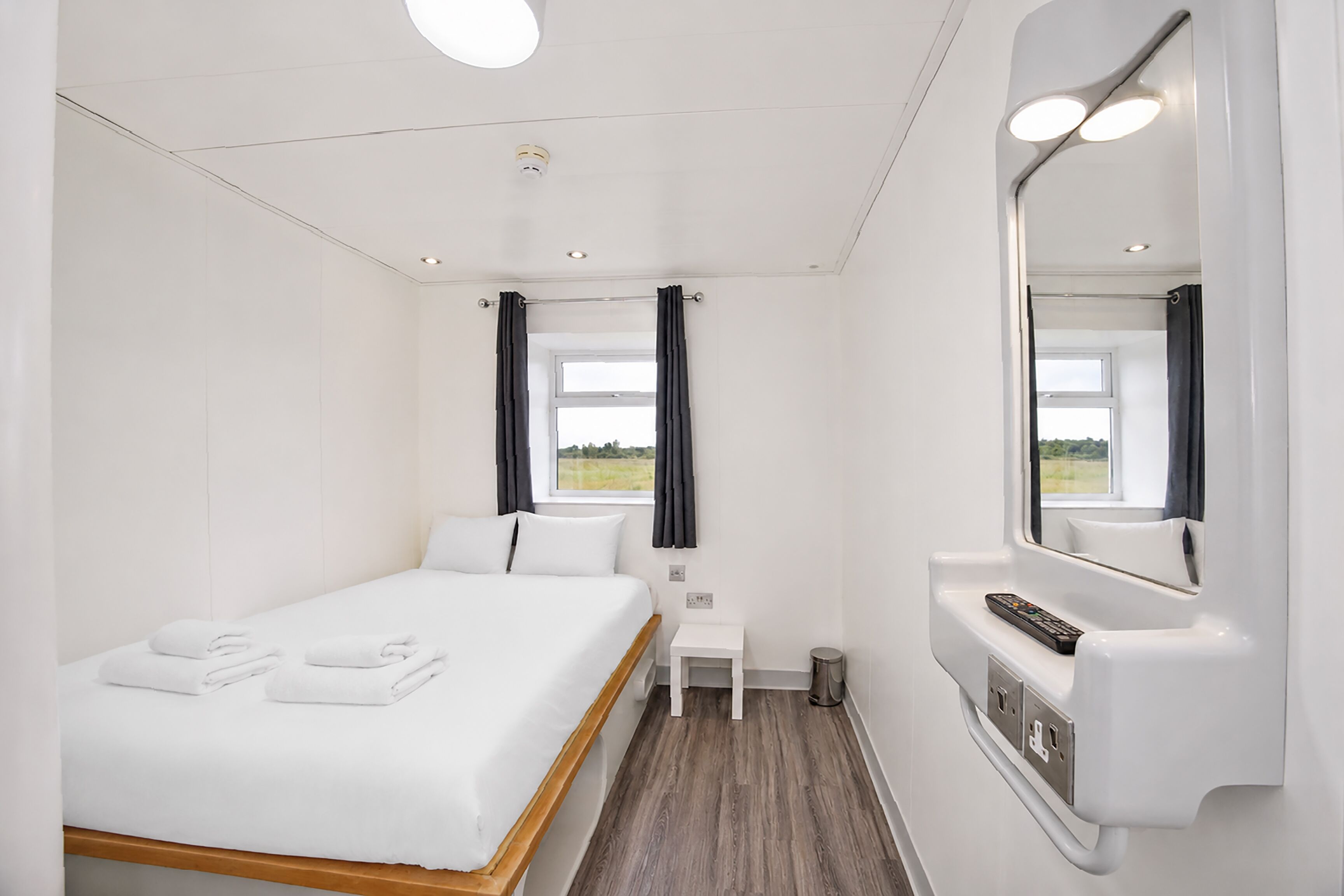 A hotel room with a bed, a small bedside table, a mirror, a window with curtains, and a view of the outdoors.
