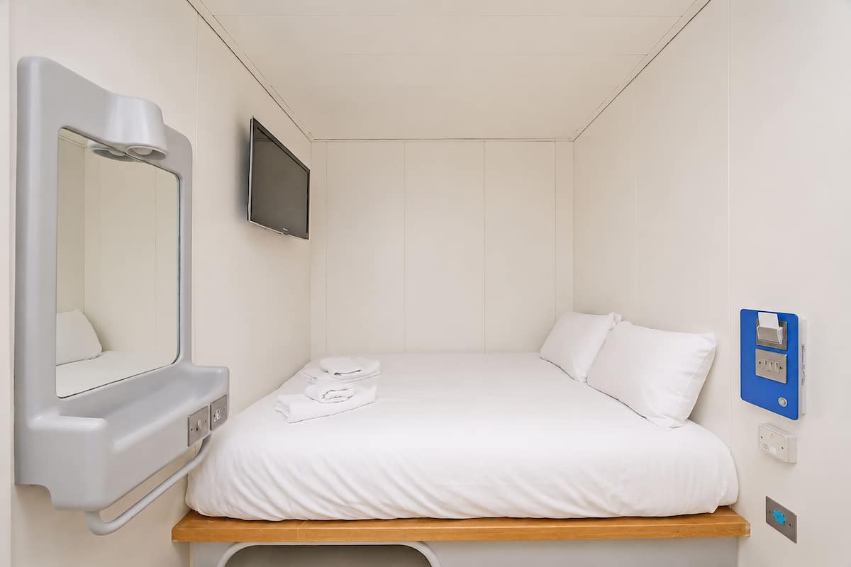 Quarto de hotel com cama, mesa de cabeceira, televisão na parede, espelho e tomada.