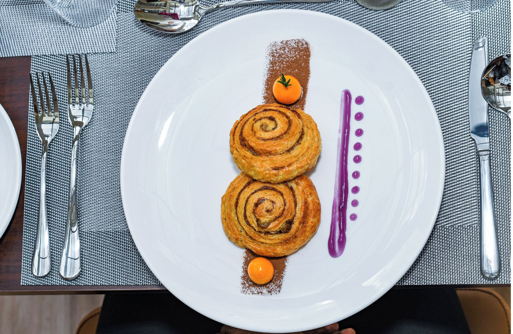 Ein Teller mit zwei spiralförmigen Gebäcken, einer kleinen orangen Frucht und einer violetten Soße.