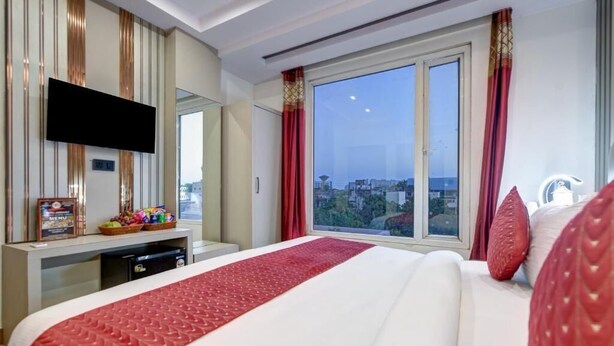 A hotel room with a large bed, a TV mounted on the wall, a desk with a basket of snacks, and a window with a city view. - Hotel Ambience (New Delhi)