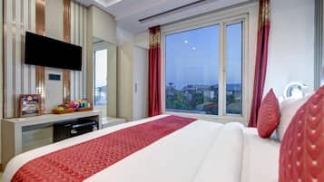 Habitación de hotel con una cama grande, un televisor montado en la pared, un escritorio con una cesta de aperitivos y una ventana con vistas a la ciudad.