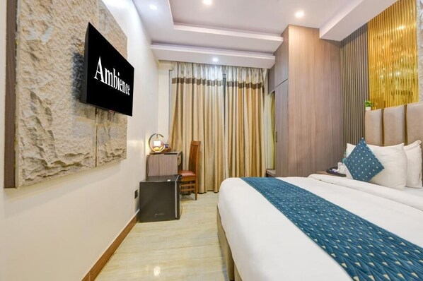 A hotel room with a bed, a TV mounted on a textured wall, a desk with a chair, and a window with curtains.