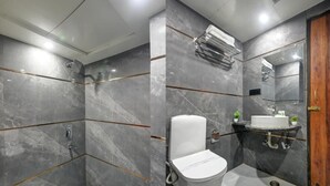 A modern bathroom with a shower, toilet, and sink, featuring marble walls and a wooden door. - Hotel Ambience (New Delhi)