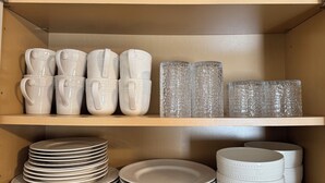 A storage area with shelves containing white ceramic mugs, clear glass tumblers, and stacked plates.