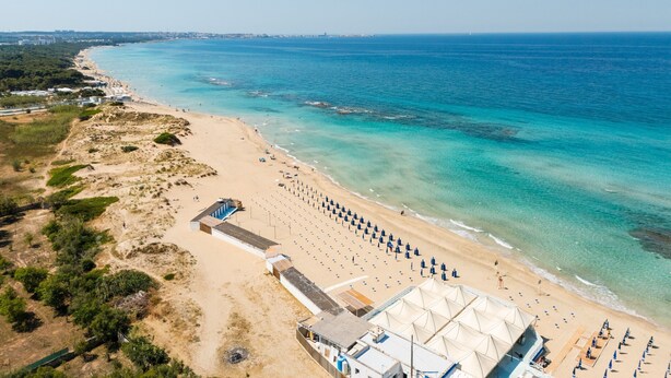 An aerial view of a beach with a large tented area, sandy beach, and clear blue water.