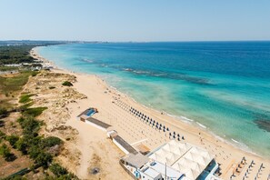 Vue aérienne d’une plage avec une grande zone abritée par des tentes, une plage de sable et une eau d’un bleu limpide.
