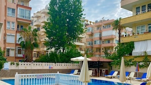A swimming pool with lounge chairs and umbrellas, surrounded by multi-story buildings. - ALANYAHOMES (Alanya)