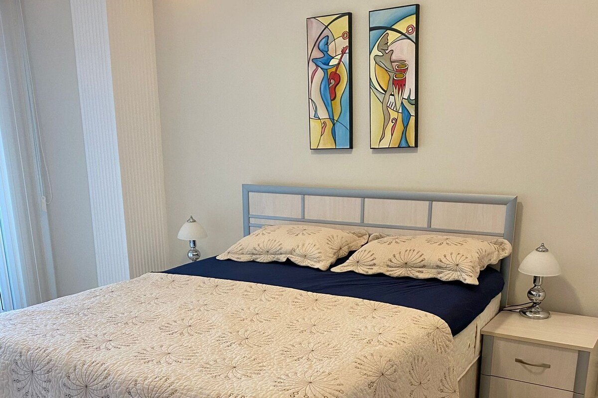 A bedroom with a bed, nightstands, a lamp, a star-patterned rug, and two abstract paintings on the wall.