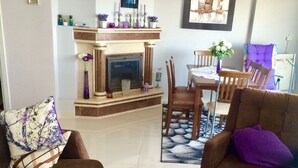 A living room with a fireplace, sofa, armchair, glass coffee table, and dining area. - ALANYAHOMES (Alanya)