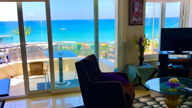 A living room with a large window overlooking the ocean, a flat-screen TV, and a dining area with a glass table. - ALANYAHOMES (Alanya)