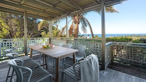 A covered outdoor seating area with a table set for a meal, overlooking a scenic view of the ocean and palm trees. - Fliptide House - ocean views, spacious living and easy access to Kiama’s beaches (Kiama)