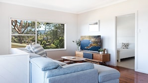 A modern living room with a sofa, TV, and a view of trees through large windows. - Fliptide House - ocean views, spacious living and easy access to Kiama’s beaches (Kiama)