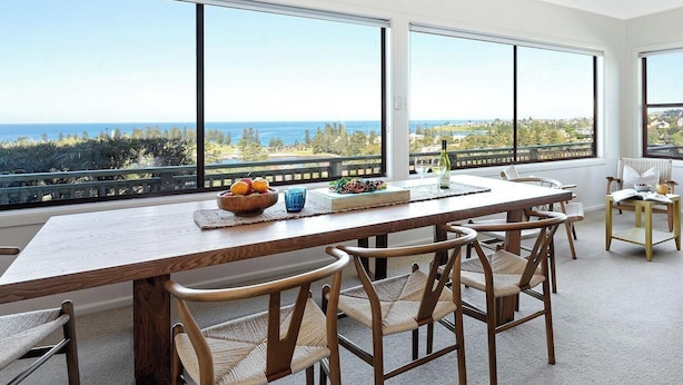 A dining area with a wooden table, wicker chairs, and a view of the ocean. - Fliptide House - ocean views, spacious living and easy access to Kiama’s beaches (Kiama)