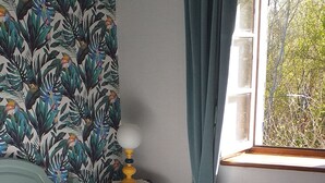 A bedroom with a bed, a nightstand with a lamp, a window with curtains, and a radiator. - MAS PROVENCAL DE CARACTERE EN PIERRE. (Goudargues)