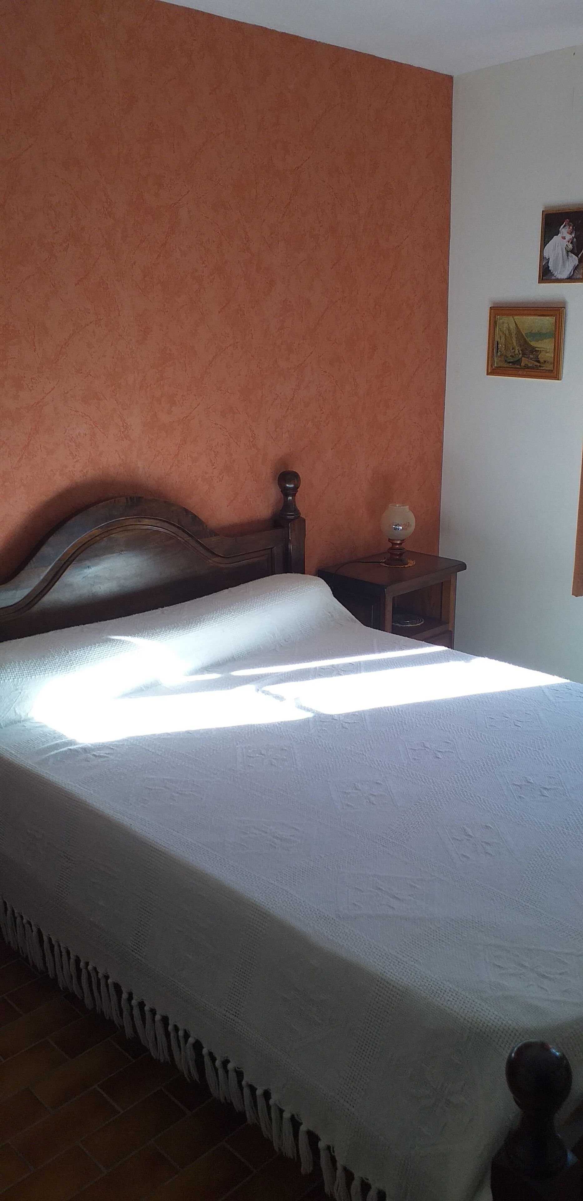 Una cama con colcha blanca, cabecera de madera, mesita de noche con lámpara y un cuadro enmarcado en la pared.