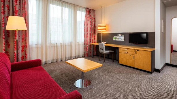 A hotel room with a red sofa, a small wooden table, a desk with a TV, and a lamp. - Sunday One-bedroom Suite Twin Beds (Neu-Isenburg)