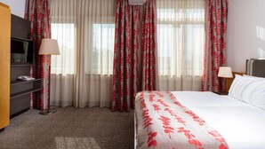 A hotel room with a bed, a desk, a television, and curtains with a floral pattern. - Sunday One-bedroom Suite Twin Beds (Neu-Isenburg)