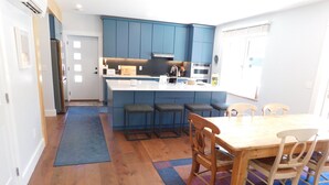 A modern kitchen with blue cabinets, a central island, bar stools, and a dining table with chairs. - Brand new GORGEOUS contemporary home (CS0054) (Wellfleet)