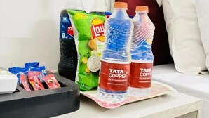 A hotel room with a bed, a nightstand, and a table with bottled water, a green snack packet, and a red and blue bottle labeled 'Tata Copper.' - Hotel RS International (New Delhi)