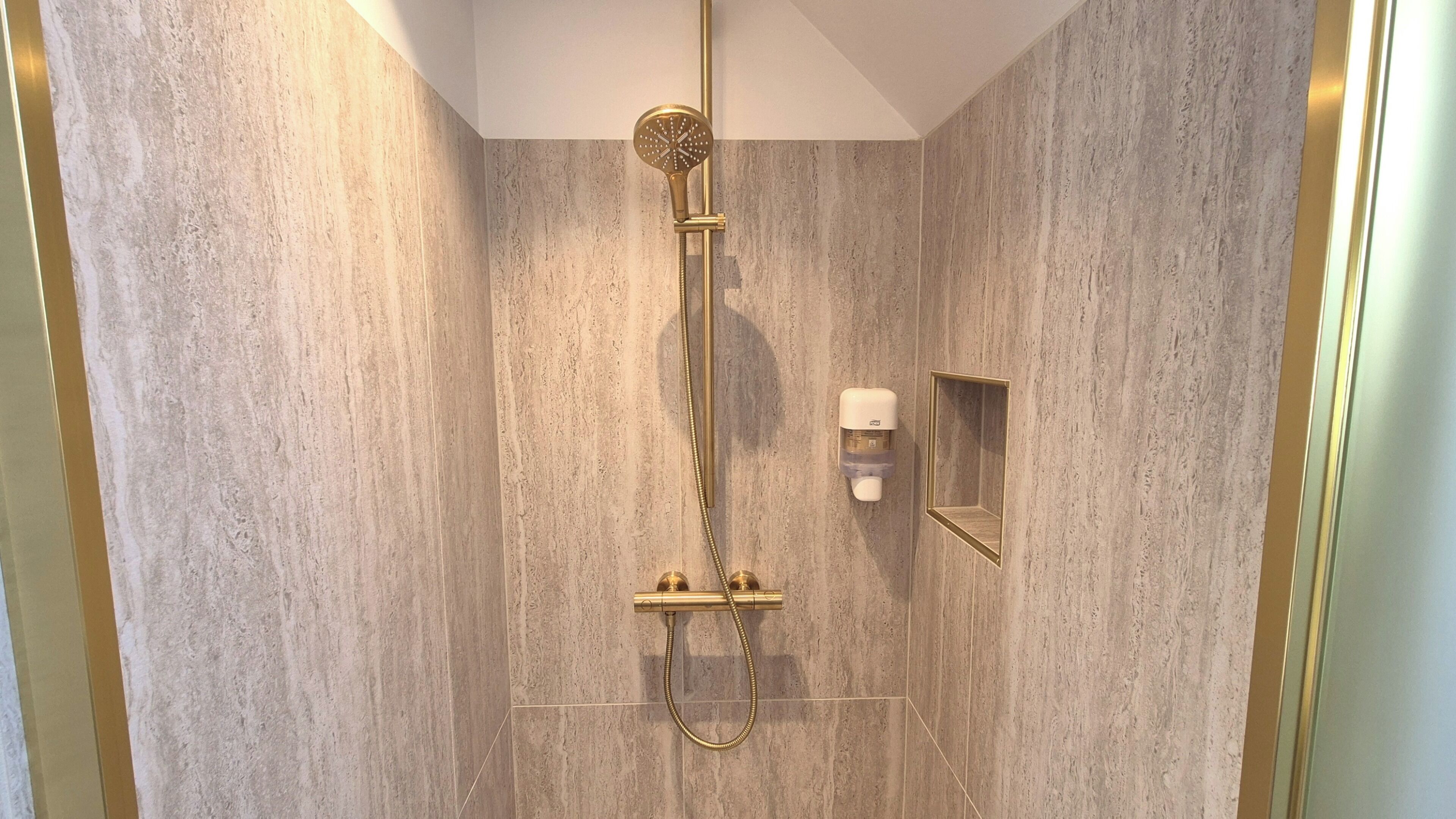 A shower with a gold showerhead and hose, a small mirror, and a soap dispenser on the wall.