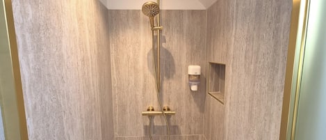A shower with a gold showerhead and hose, a small mirror, and a soap dispenser on the wall.