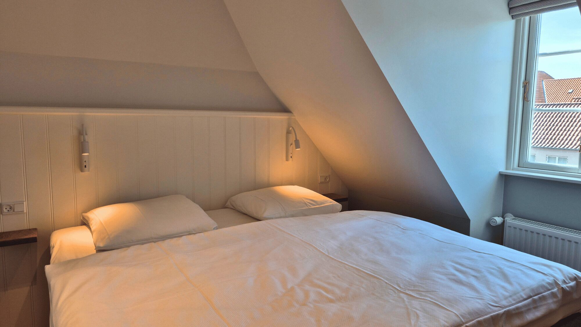 A bedroom with a sloped ceiling, a bed with white bedding, a nightstand, a wall-mounted lamp, and a window with a view of buildings.