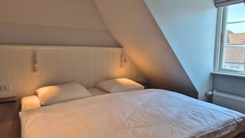 Un dormitorio con techo inclinado, cama con ropa de cama blanca, mesita de noche, lámpara de pared y ventana con vistas a edificios.
