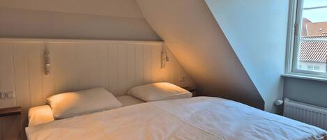 A bedroom with a sloped ceiling, a bed with white bedding, a nightstand, a wall-mounted lamp, and a window with a view of buildings.