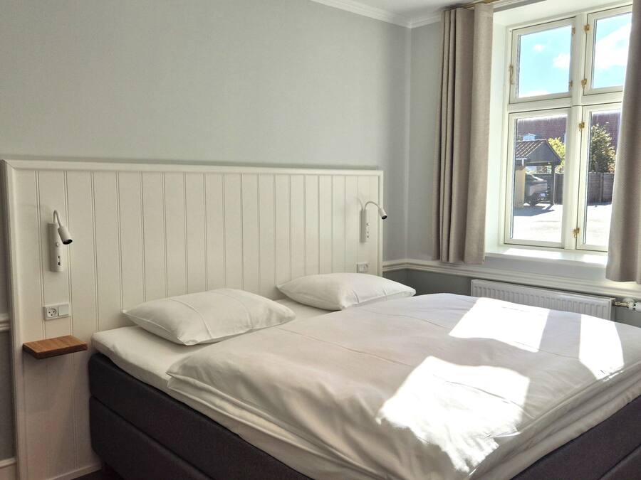 A bedroom with a bed, a window with curtains, a bedside table, and a wall-mounted light fixture.
