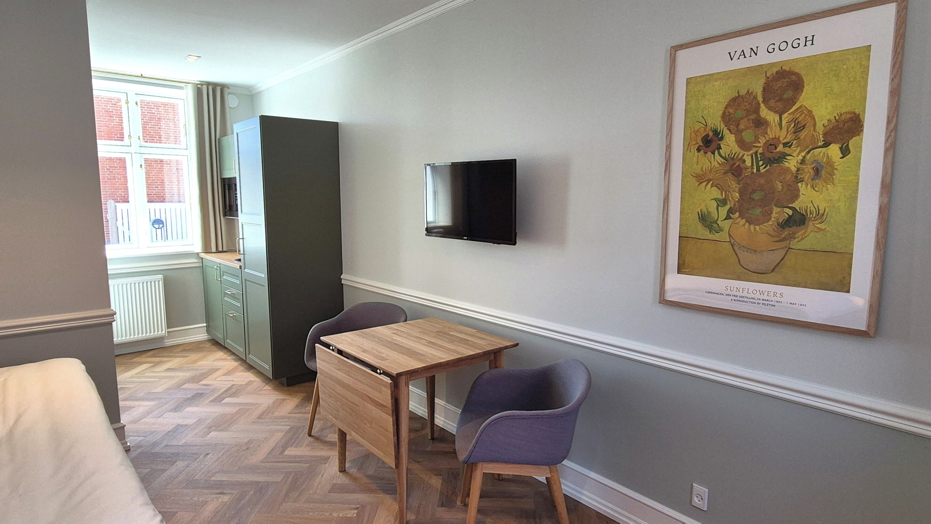 Une pièce comprenant une table en bois, deux chaises, un téléviseur fixé au mur et une œuvre d’art encadrée représentant des tournesols.