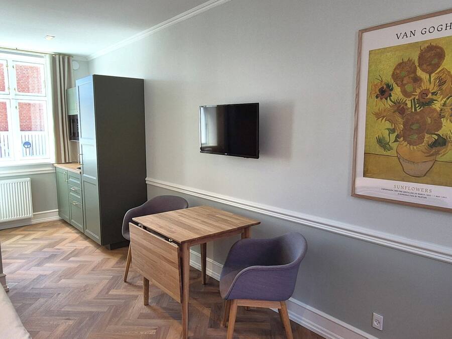 Una habitación con una mesa de madera, dos sillas, un televisor montado en la pared y una obra de arte con girasoles enmarcados.