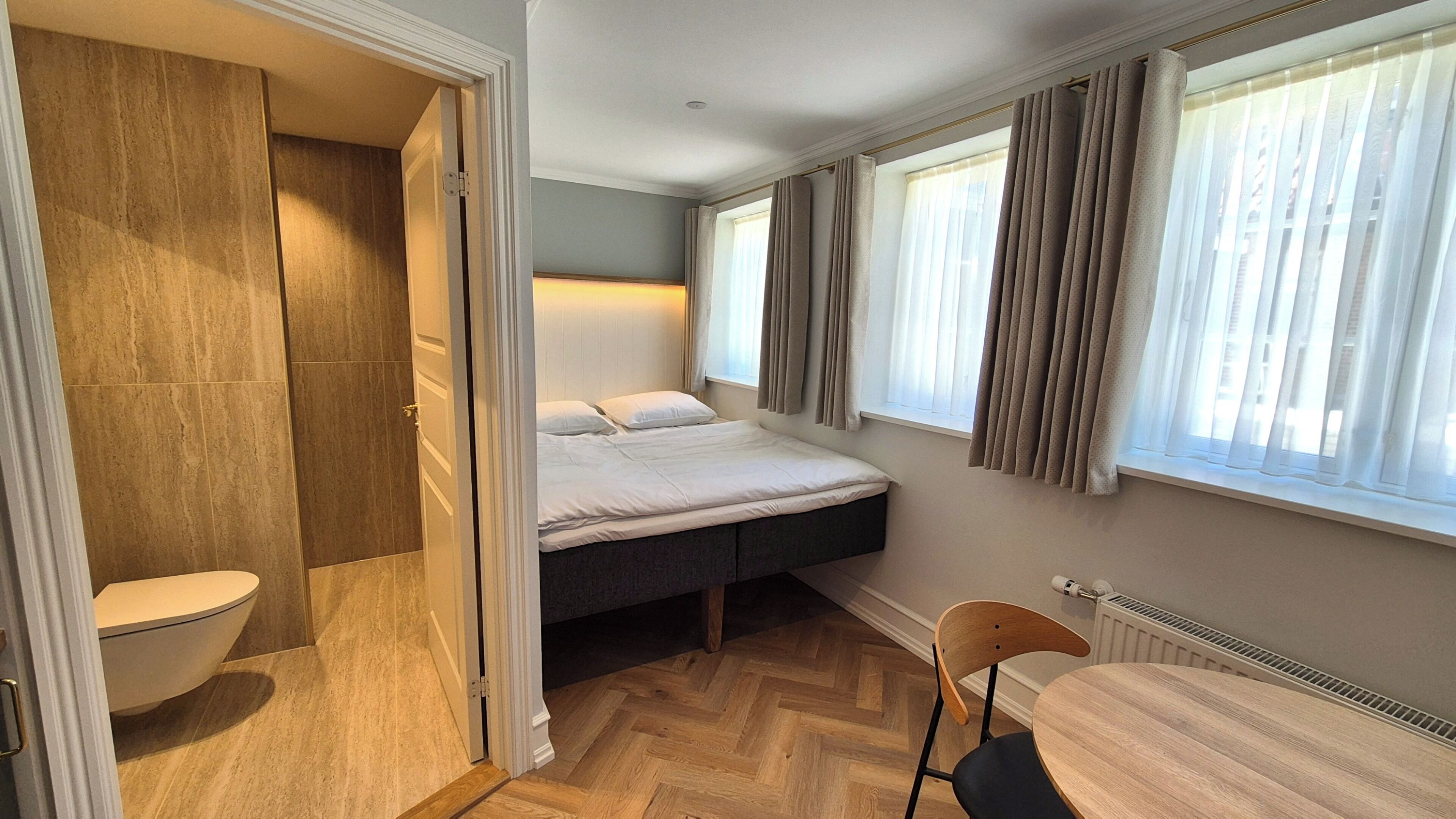 A compact room with a bed, a small table, a chair, a toilet, and a window with curtains.