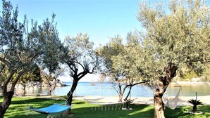 A hammock between trees, a grassy area, and a body of water in the background. - Private Beachfront House Irene Mare Vagionia Bay (Poros)