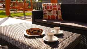 A wicker outdoor seating area with a wooden swing set, a cushioned sofa with a patterned pillow, and a table with a plate of food and two cups. - Chalet Mountain View Nova Lesna (Nová Lesná)