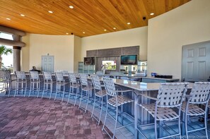 Zona de bar con sillas metálicas y mostrador, espacio de cocina con televisor, y una puerta.